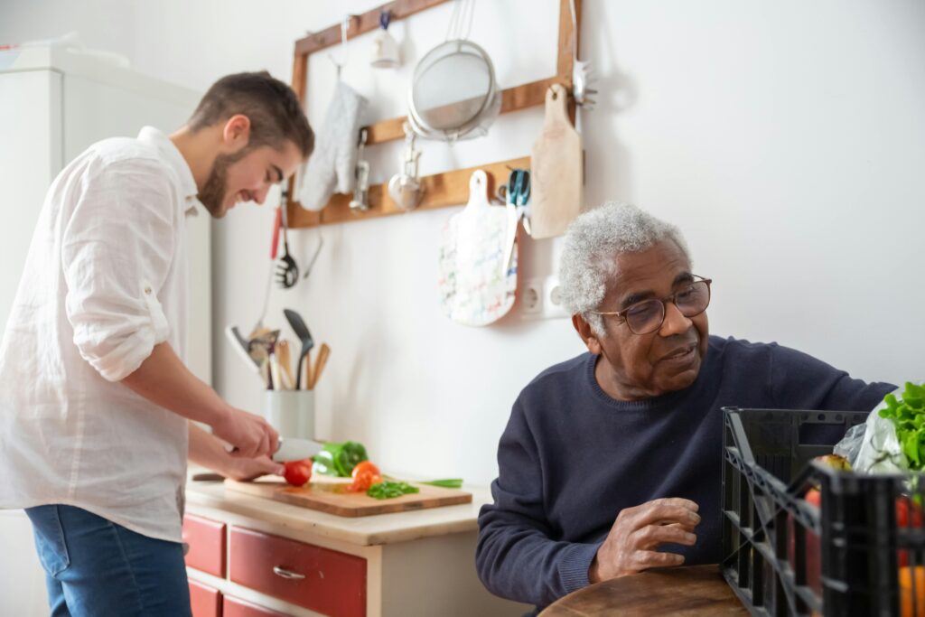 A live-in caregiver prepares a meal as he chats with a senior man